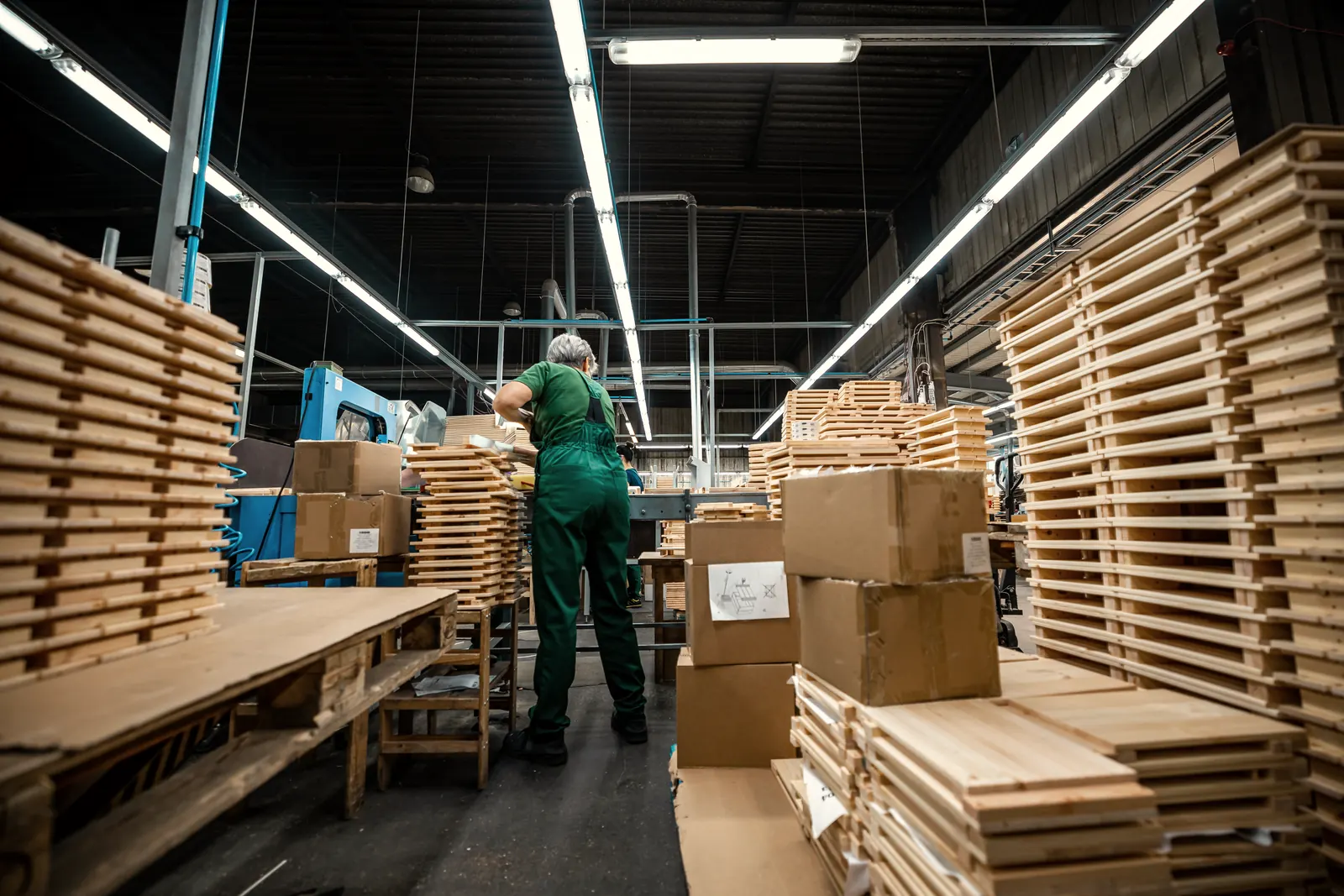 Un ouvrier aux cheveux gris, en salopette verte, travaille de dos entre des piles de palettes et de boîtes, dans une usine de bois éclairée aux néons